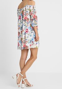 Květinové šaty bez ramínek s třičtvrtinovými rukávy, s výraznými vícebarevnými vzory na bílém pozadí. Spárováno s bílými vysokými sandály.