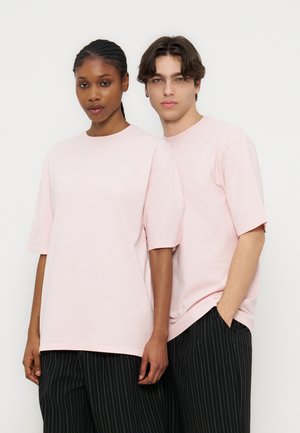Deux jeunes adultes debout côte à côte, portant des t-shirts oversize rose clair et des pantalons noirs à fines rayures, sur un fond blanc uni.