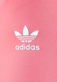 Růžová bavlněná látka s bílým logem Adidas se třemi proužky a symbolem trojlístku, umístěná uprostřed spodní části.