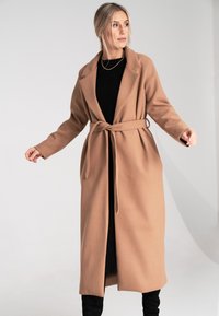 Kamelowy płaszcz o długim kroju z paskiem w talii, z wyłogowym kołnierzem i długimi rękawami, noszony na czarnej sukience z prążkami i zestawiony z czarnymi botkami.