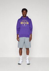 Lila pulóver "LAKERS" logóval, szürke rövidnadrág, fehér sportcipő, egyszínű háttéren állva. Hétköznapi sportos viselet, letisztult dizájn.