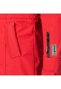 Chaqueta roja con tejido texturizado, múltiples bolsillos y cremallera. Incluye una etiqueta de marca y detalles de herrajes en contraste.