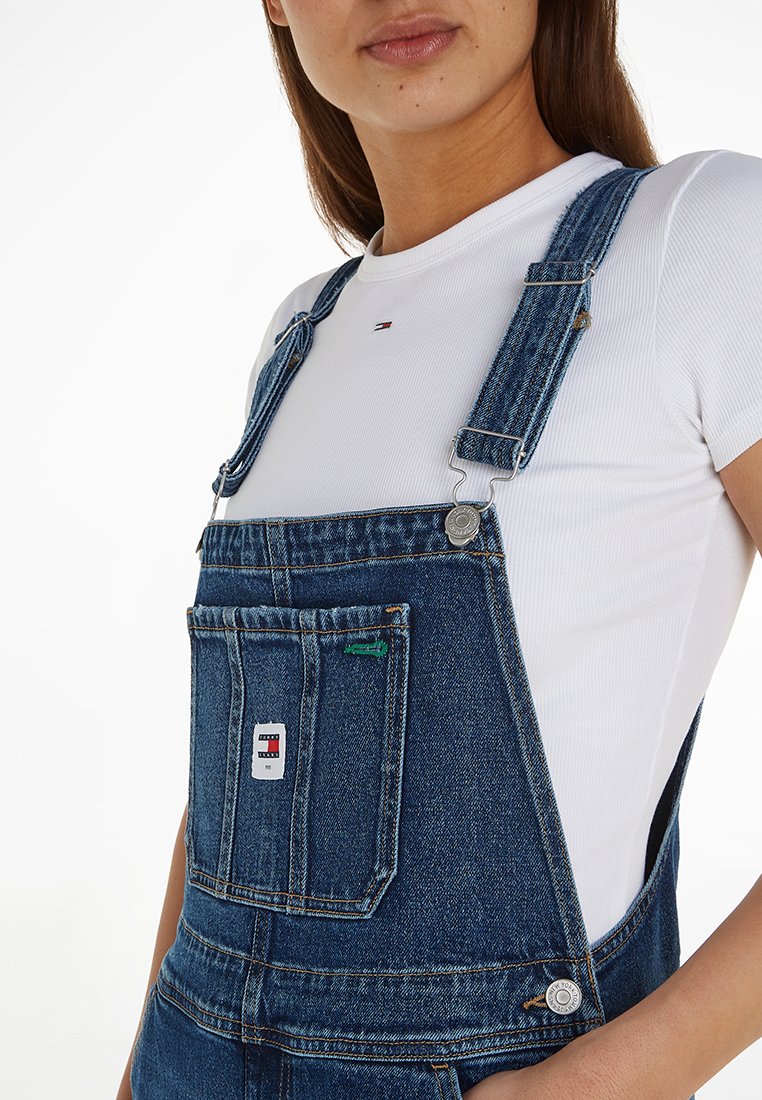 Salopette en denim bleu foncé avec bretelles ajustables, boutons en métal, poche avant et patch logo sur le devant. Portée par-dessus un t-shirt blanc.