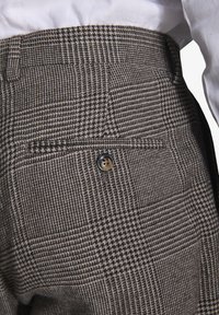 Brune og grå houndstooth-mønstrede bukser med en sort knap, sidelomme og en tekstureret stofoverflade. Skræddersyet design.