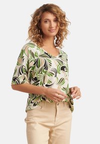 Kurzärmlige Bluse in Weiß mit grünen, schwarzen und beigen Blattmustern. V-Ausschnitt, lockere Passform, weiche Textur, kombiniert mit hellen Hosen.
