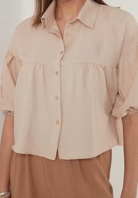 Jasno beżowa krótka bluzka z kołnierzykiem, bufkami, zapięciem na guziki i marszczeniem w okolicy biustu, wykonana z miękkiej, teksturowanej tkaniny.