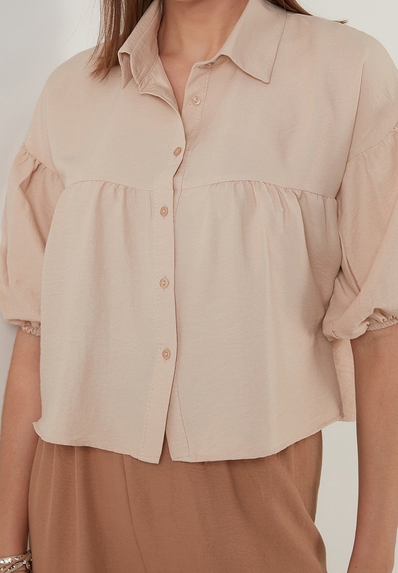 Jasno beżowa krótka bluzka z kołnierzykiem, bufkami, zapięciem na guziki i marszczeniem w okolicy biustu, wykonana z miękkiej, teksturowanej tkaniny.