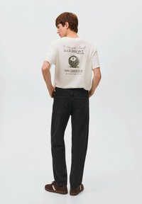 Camiseta blanca con estampado gráfico que presenta una bola ocho y texto. Vaqueros de mezclilla oscura, zapatos marrones con acentos blancos. Estilo y corte informal.