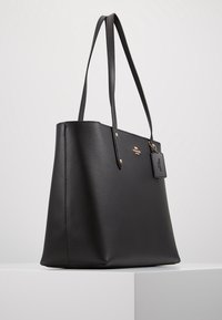 Musta nahkainen käsilaukku, jossa on teksturoitu pinta, pitkät hihnat, logokoriste ja irrotettava merkki. Yksinkertainen, suorakulmainen muoto.