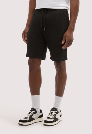 Uomo con pantaloncini neri con coulisse, calzini bianchi alla caviglia e scarpe da ginnastica bianche e nere in piedi davanti a uno sfondo semplice.