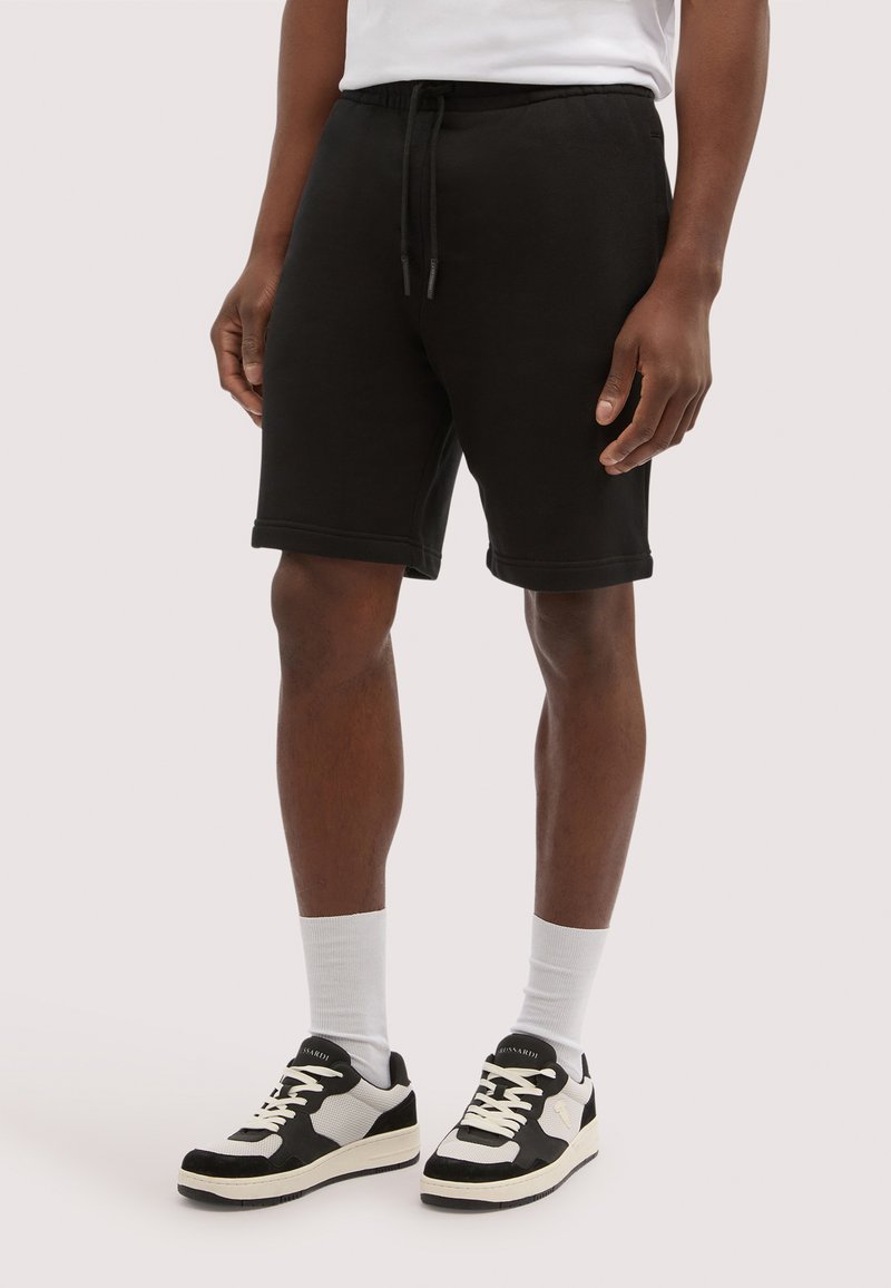 Mężczyzna ubrany w czarne spodenki ze sznurkiem, białe skarpetki za kostkę oraz czarno-białe sneakersy, stojący na jednolitym tle.