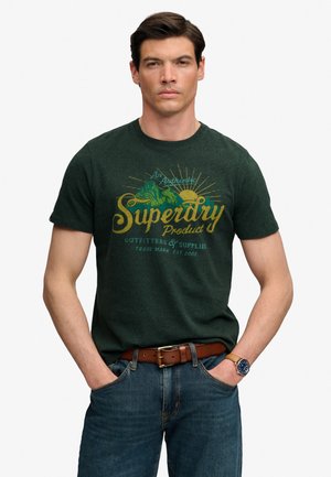 Homme aux cheveux foncés porte un t-shirt vert Superdry, un jean bleu, une ceinture en cuir marron et une montre-bracelet, debout les mains dans les poches.
