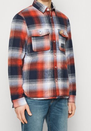 Koszula w czerwono-biało-granatową kratę z guzikami i przednimi kieszeniami na klapę, noszona przez osobę w niebieskich dżinsach na jednolitym tle.