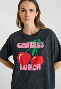 Mujer con una camiseta negra con grandes cerezas rojas y el texto rosa "AMANTE DE LAS CEREZAS" en el frente, de pie contra un fondo liso.