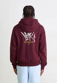 Bordeaux hoodie med snor, der har hvid grafisk tekst og logo på ryggen. Ribbet manchetter og kant. Båret med lyseblå bukser.