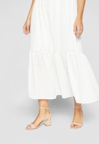 White midi dress with eyelet pattern, tiered hem, and short sleeves. Pair of beige block-heel sandals with ankle strap and metallic accent.