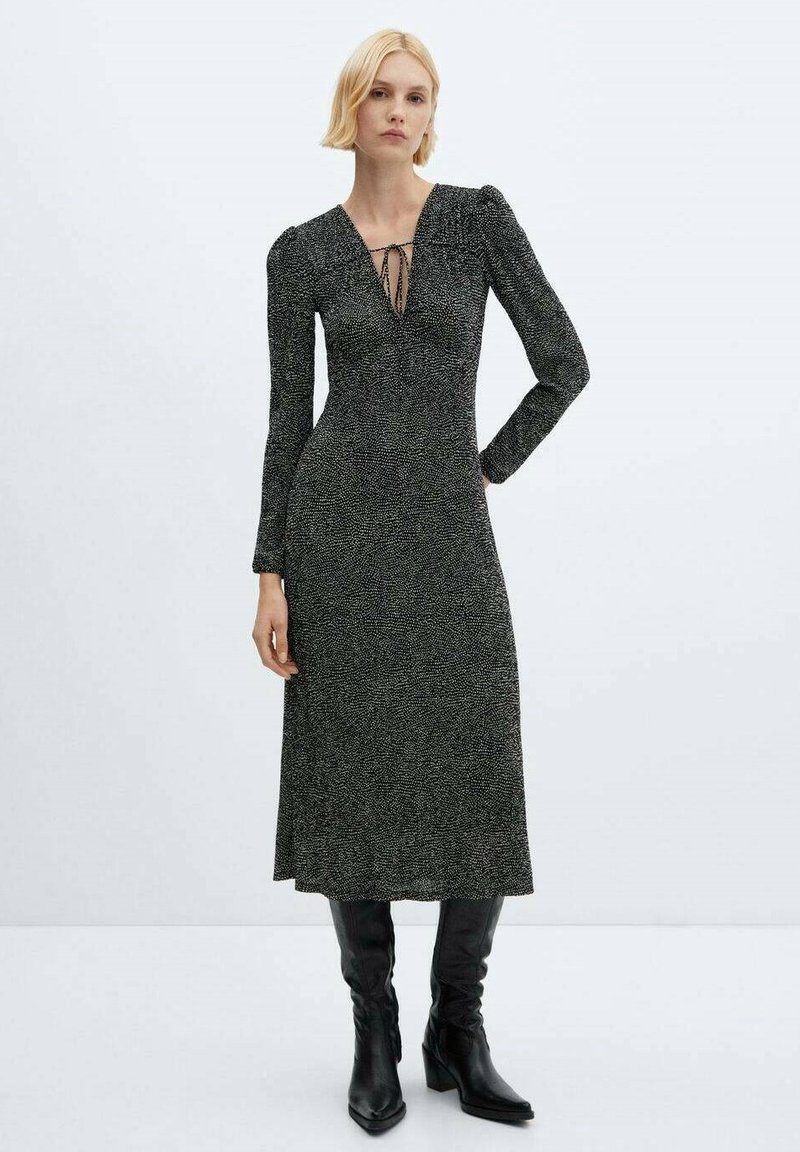 Abito nero a maniche lunghe con finitura testurizzata, scollatura a V profonda e dettagli con fiocco sul davanti, che arriva a metà polpaccio. Indossato con stivali neri al ginocchio.