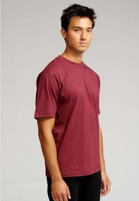 Ung mand med kort, mørkt hår, iført en ensfarvet maroon t-shirt med korte ærmer og sorte bukser, stående foran en lys baggrund.