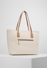 Torba typu tote w jasnym kremowym skórzanym wykończeniu w drobne prążki, z dwoma brązowymi uchwytami oraz przednią kieszenią na zamek; prostokątny kształt; gładka tekstura.