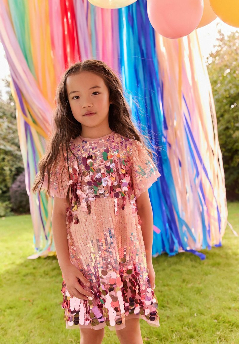 Ragazza giovane con capelli lunghi indossa un vestito rosa con paillettes, in piedi sull’erba con decorazioni colorate di arcobaleno e palloncini dietro di lei.