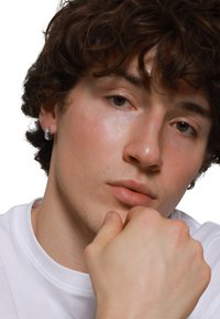 Orecchino a cerchio d'argento su un giovane con capelli ricci castani, che indossa una maglietta bianca, appoggiando il mento sulla mano, su uno sfondo neutro.