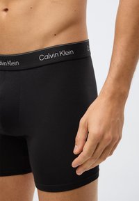 Close-up van een man in zwarte Calvin Klein boxershorts met de zichtbare merkband, toont torso en rechterhand.