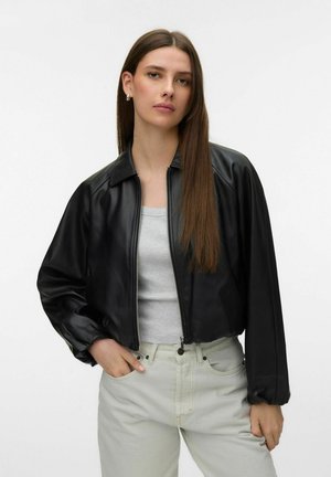 Ung kvinde med langt brunt hår iført en sort lynlåsjakke, lysegrå skjorte og lyse jeans, stående med den ene hånd i lommen.