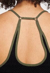 Närbild av en bakvy som visar en hudtonad bakgrund och två olivgröna remmar som kopplas samman vid en silverring på ett svart strukturerat tyg.