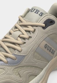 Κοντινή λήψη μπεζ και γκρι αθλητικού παπουτσιού Guess που δείχνει υφή υφάσματος, λεπτομερείς ραφές, κορδόνια και το όνομα της μάρκας στη γλώσσα και στο πλάι.