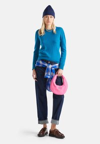 Modrý pletený svetr, kostkovaná košile uvázaná v pase, tmavé džínové kalhoty a hnědé boty. Růžová kulatá taška s dekorativním logem.