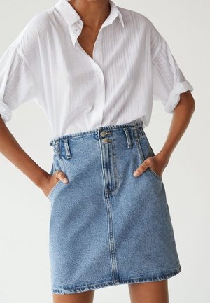 Personne portant un chemisier blanc à manches retroussées, rentré dans une jupe en denim bleu taille haute avec des poches avant et des boutons doubles.