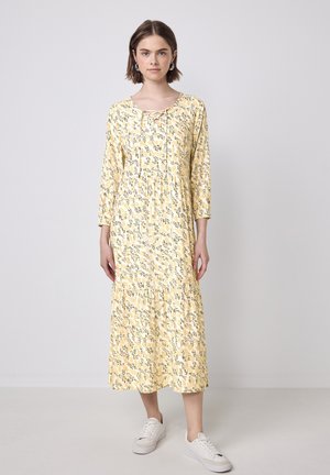 Robe florale jaune avec des manches 3/4, taille froncée et ourlet en couches. Le tissu présente un motif de feuilles ; le mannequin porte des baskets blanches.