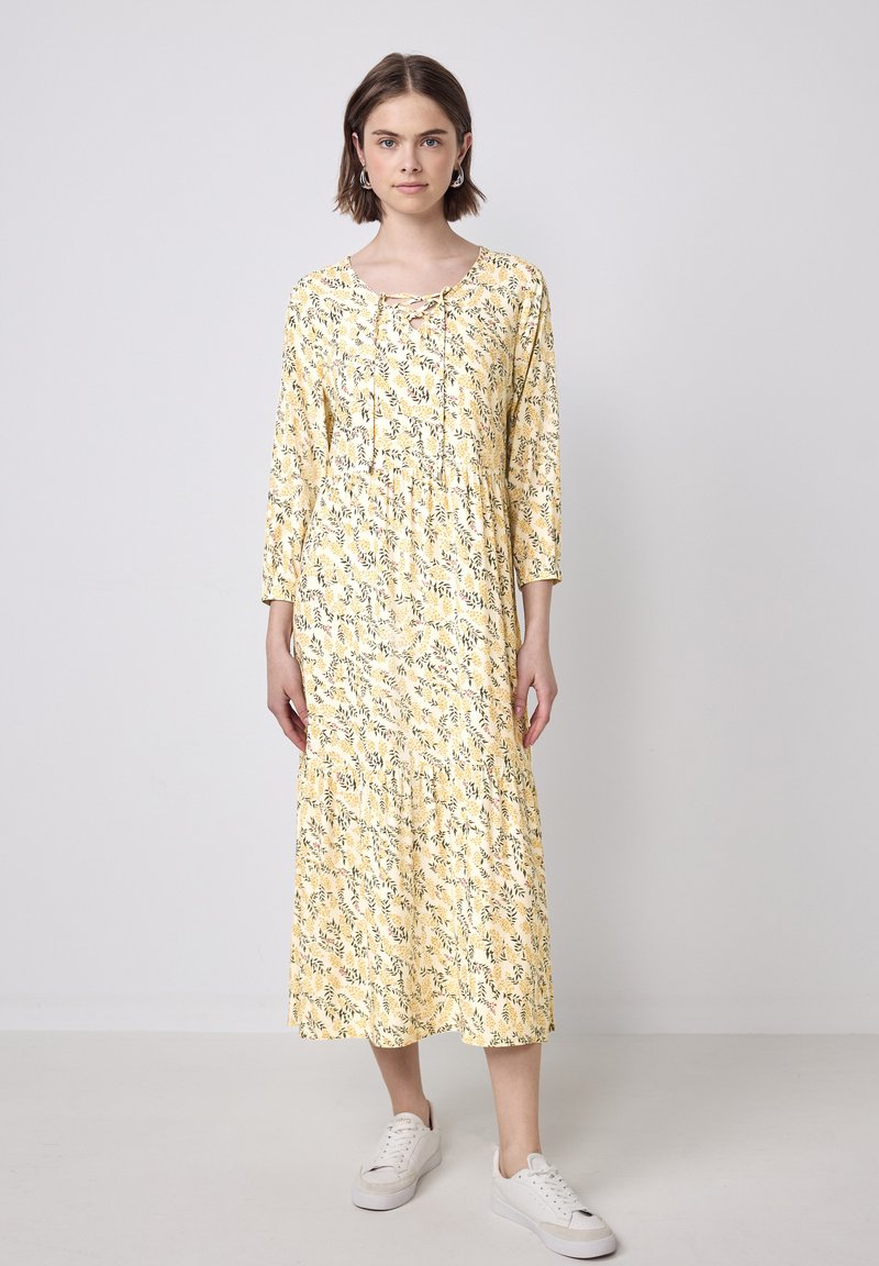 Gelbes Blumenkleid mit 3/4 Ärmeln, geraffter Taille und mehrlagigem Saum. Der Stoff zeigt ein Blattmuster; das Model trägt weiße Sneakers.