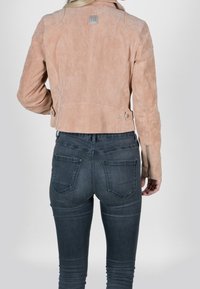 Sömsuede, ljusrosa kort jacka med spetsig krage, sidobältesdetaljer och dragkedjor på ärmarna, i kombination med mörka jeans.