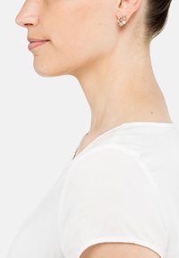 Goldohrringe mit floralem Design, die mit Perlenakzenten verziert sind. Das Modell trägt ein einfaches weißes T-Shirt. Die Profilansicht hebt die Details der Ohrringe hervor.