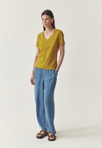 Sinepikollase V-kaelusega T-särk koos lahtiste siniste teksapükstega. Modell kannab pruune sandaale. Minimalistlik disain lühikeste varrukatega ja vaba istuvusega.