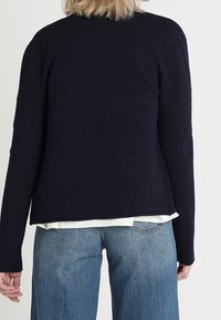 Námořnický pletený svetr s dlouhými rukávy, kulatým výstřihem a uvolněným střihem. Vyznačuje se texturovaným povrchem a asymetrickým lemem.