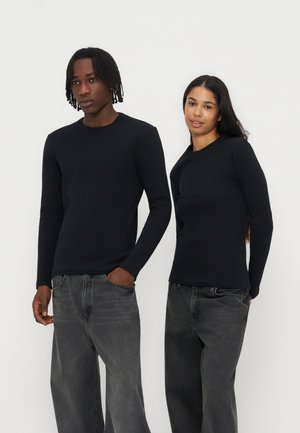 Deux adultes portant des hauts noirs à manches longues et des jeans gris se tiennent devant un fond blanc uni, face à l'avant avec des poses détendues.