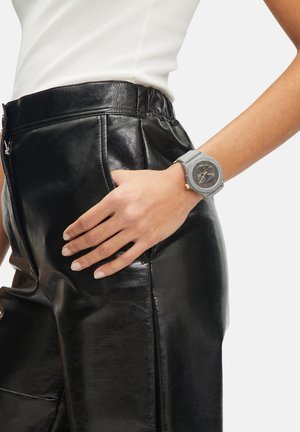 Grå ur med en rund urskive, guldfarvede detaljer og en silikone rem. Hånd hviler på et sort kunstlæderbeklædning med en skinnende overflade.