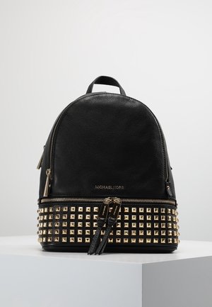 Zwarte Michael Kors leren rugzak met goudkleurige studs langs de onderkant en dubbele ritstrekkers op een witte ondergrond.