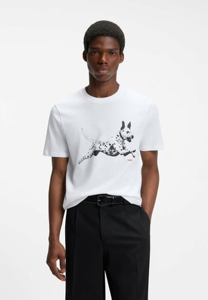 Hombre con una camiseta blanca con un gráfico de perro saltando en blanco y negro, cinturón negro y pantalones negros sobre un fondo blanco liso.