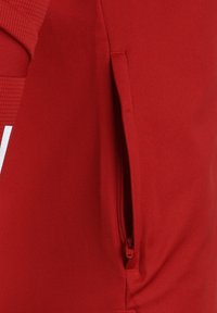 Rote Sportbekleidung aus einem strukturierten Stoff, mit einer seitlichen Reißverschlusstasche und kontrastierender weißer Markenkennzeichnung.