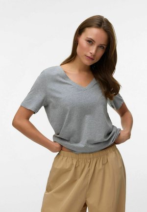 Woman with long brown hair wearing a gray V-neck t-shirt and beige elastic waist pants, hands resting on hips against a white background.