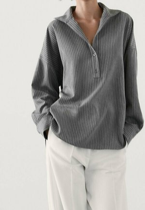 Woman wearing a loose, long-sleeve, gray ribbed shirt with buttons and white high-waist pants, hands in pockets, against a plain background.