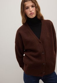 Brauner, knöpfbarer Cardigan aus weichem, strukturiertem Stoff; verfügt über einen tiefen V-Ausschnitt und gerippte Bündchen. Wird über einem schwarzen Rollkragenpullover getragen.