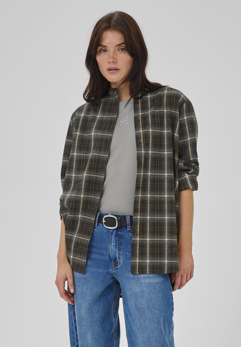 Kvinde iført en åbenstående mørk ternet skjorte over en grå top, blå højtaljede jeans med et sort bælte, stående mod en grå baggrund.