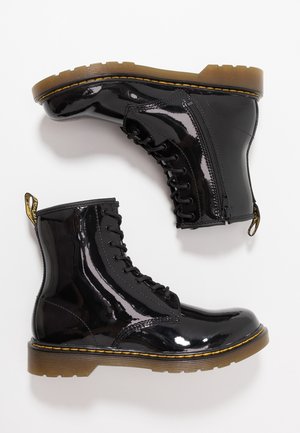 Paire de bottines noires brillantes à lacets avec fermetures éclair latérales et semelles épaisses marron avec surpiqûres jaunes sur fond blanc.