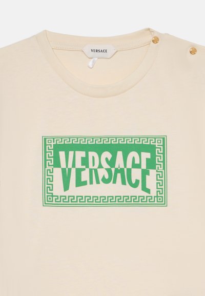 Μπεζ μπλουζάκι Versace με πράσινο γεωμετρικό περίγραμμα και μεγάλο πράσινο λογότυπο Versace εμπρός, με χρυσά κουμπιά στον ώμο.