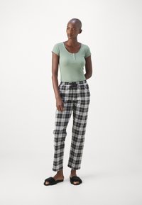 Grön ribbad topp med kort ärm och knappad halsringning, tillsammans med svart- och vitrutiga pyjamasbyxor och svarta mjuka slidesandaler.