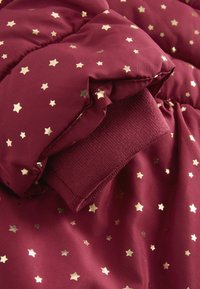 Chaqueta acolchada burdeos con patrón de estrellas doradas, con puños acanalados. Textura suave y brillante con secciones cosidas para aislamiento.